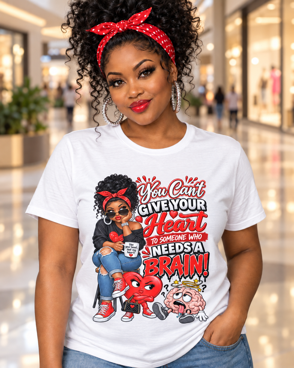 A woman wearing a white graphic t-shirt with a red and black cartoon design and the text 'You Can't Give Your Heart to Someone Who Needs a BRAIN!'. She is posing in a brightly lit indoor mall environment.